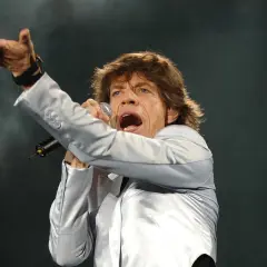 Mick Jagger śpiewa, wykonując swoje legendarne utwory. Wokalista w srebrnej marynarce z mikrofonem w dłoni.