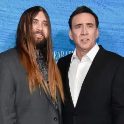 Weston Coppola Cage z długimi włosami i Nicolas Cage w czarnym garniturze pozują do zdjęcia na niebieskim tle.