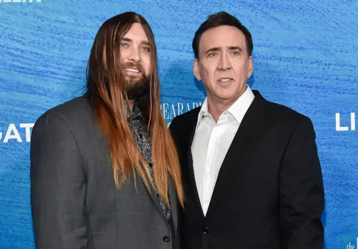 Weston Coppola Cage z długimi włosami i Nicolas Cage w czarnym garniturze pozują do zdjęcia na niebieskim tle.