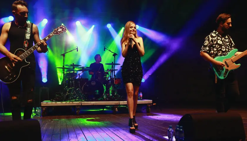 Zespół Łzy skład na scenie, wokalistka w błyszczącej sukience, gitarzysta i basista grają, perkusista w tle.