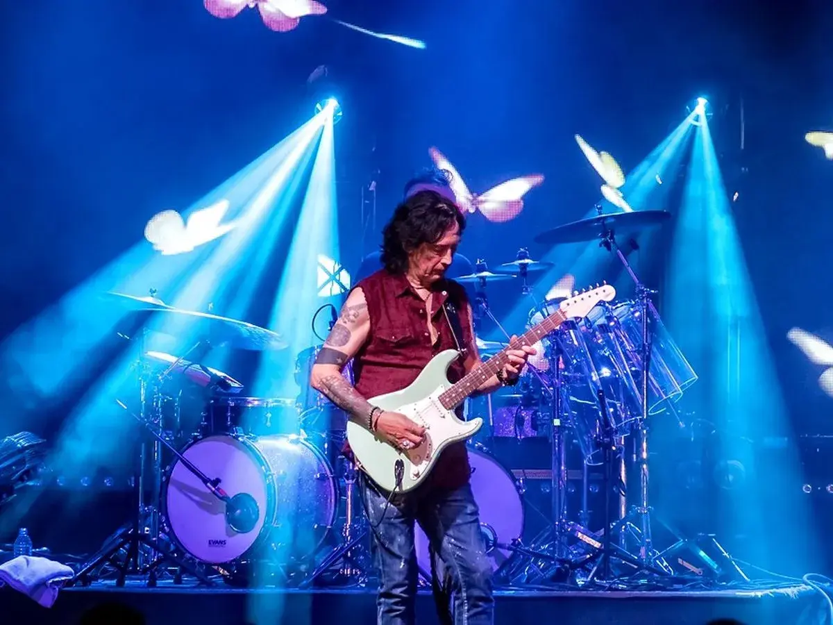 Mężczyzna gra na gitarze elektrycznej na scenie, w tle perkusja i niebieskie światła z motylami.