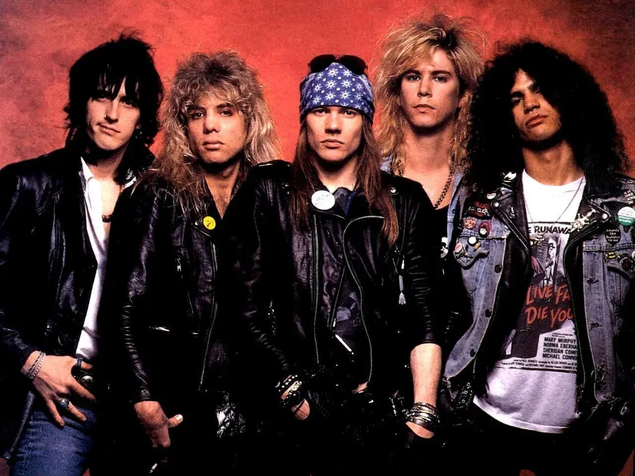 Guns N' Roses członkowie w skórzanych kurtkach i z charakterystycznymi fryzurami. Axl Rose w bandanie, Slash z burzą loków.