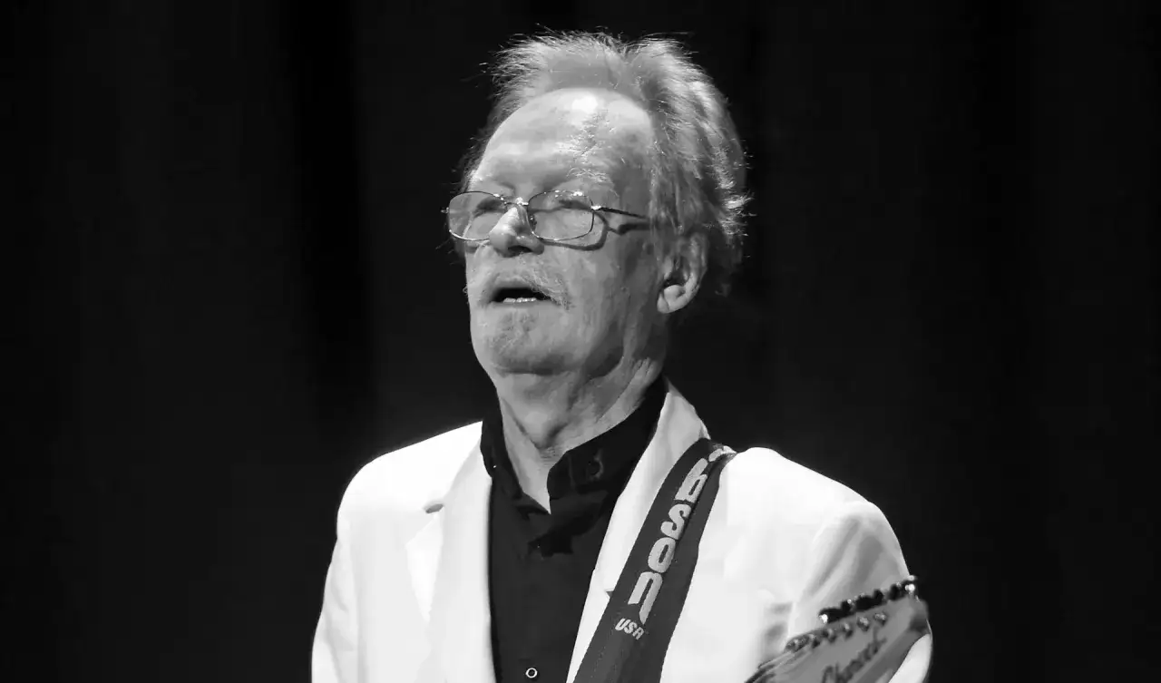 Starszy mężczyzna w białej marynarce i czarnej koszuli, z okularami na nosie, gra na gitarze. Na pasku gitary widnieje napis 