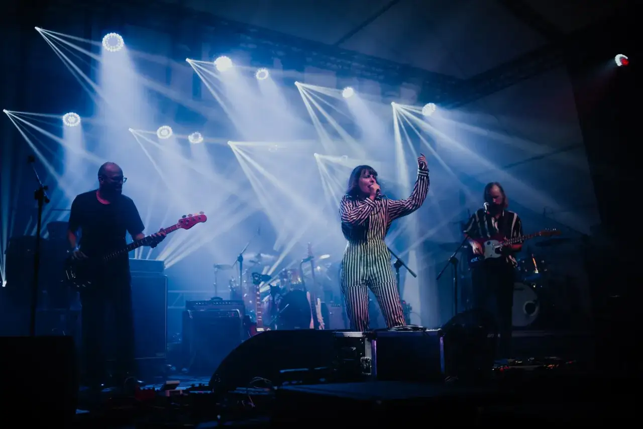 Izzy i The Black Trees na scenie, otoczeni niebieskimi światłami. Wokalistka w pasiastym stroju, gitarzyści i perkusja.