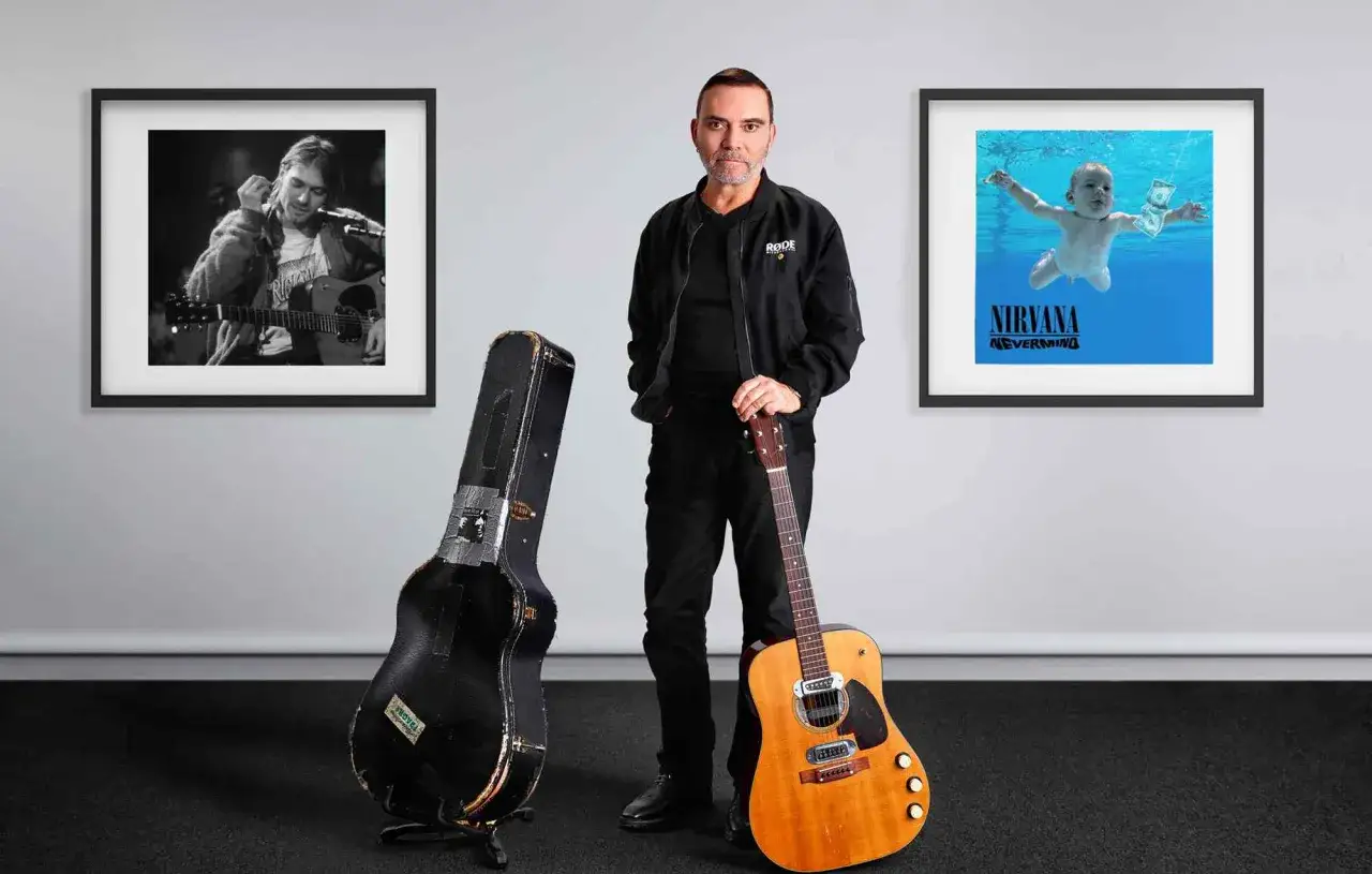 Mężczyzna z gitarą i futerałem, obok obrazów z Kurtem Cobainem i okładką