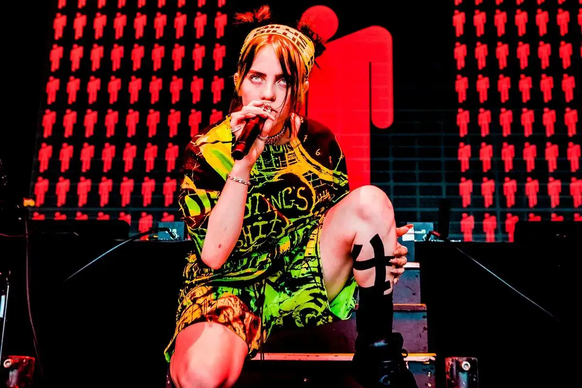Billie Eilish w Polsce na koncercie, w neonowym stroju, z mikrofonem w dłoni. Tło zdobią czerwone sylwetki ludzi.